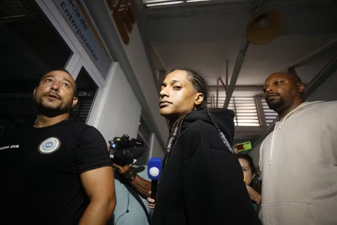 Rapper Oruam, de tranças e moletom preto, acompanhado por policial civil com emblema da corporação e outro homem de camisa bege em delegacia