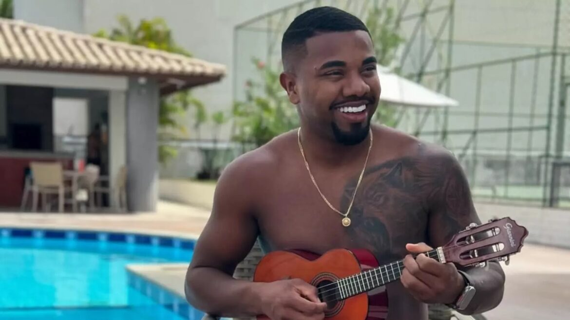 Davi Brito sorrindo, sem camisa, toca violão marrom, com piscina azul ao fundo