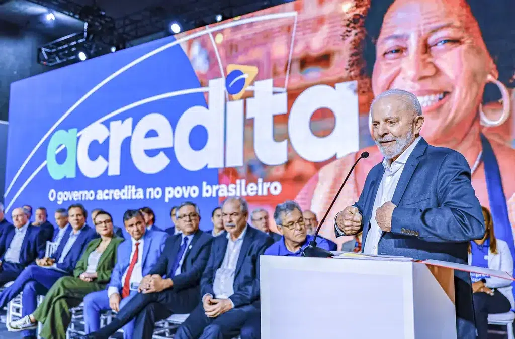 Programa Acredita: microcrédito facilitado para famílias do CadÚnico; saiba como adquirir 6 Presidente Lula em púlpito branco discursando no evento Acredita, com ministros sentados ao fundo e painel azul