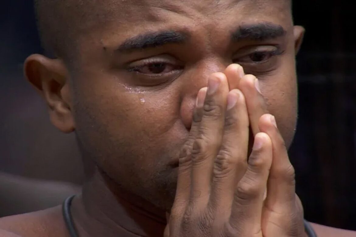 Homem com expressão emocionada, chorando, mãos cobrindo parcialmente o rosto, fundo escuro