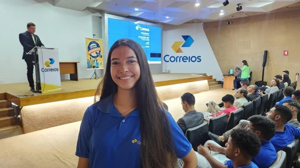 Últimas horas! Inscrições para 548 vagas nos Correios terminam hoje 6 Jovem de cabelos longos e camiseta azul dos Correios sorri em auditório durante evento do programa Jovem Aprendiz