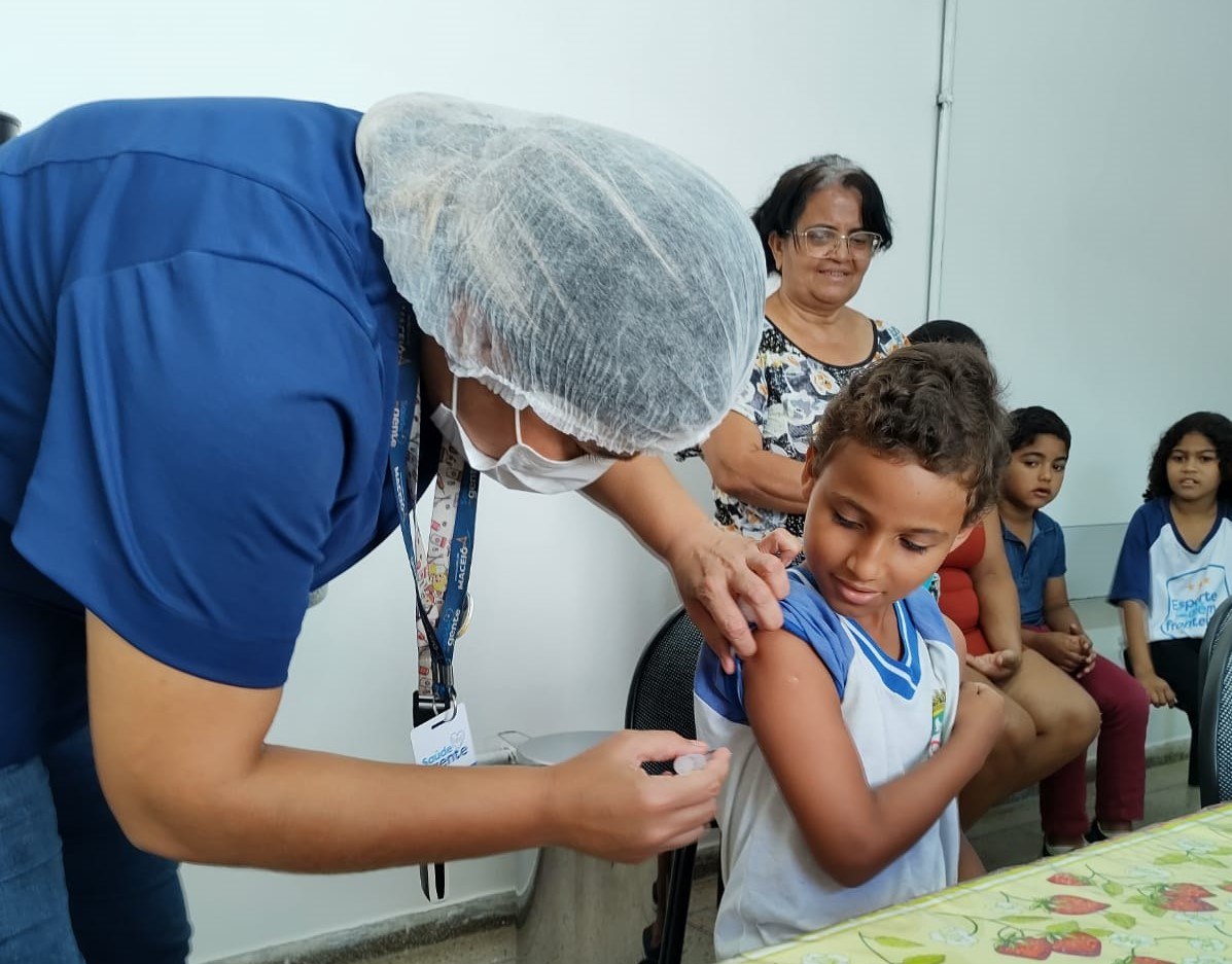 Vacinação nas escolas continua até o dia 30; veja quem precisa se vacinar 6 Profissional de saúde com touca e máscara aplica vacina em braço de menino com cabelos cacheados e uniforme escolar azul