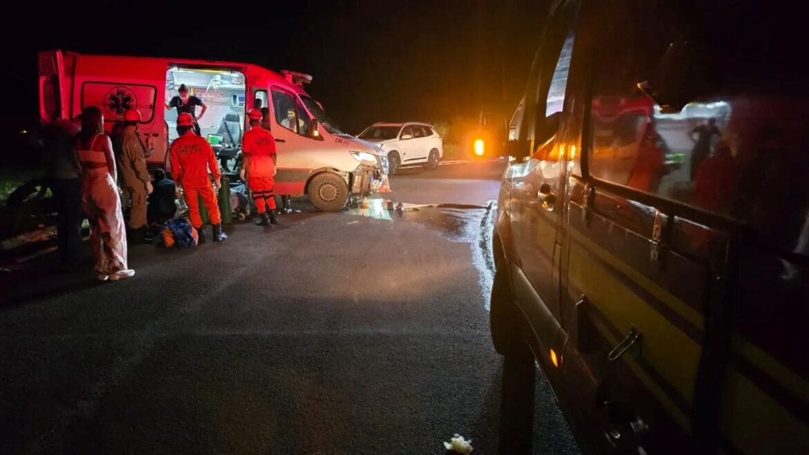 Cena noturna de acidente em rodovia mostrando uma ambulância branca com a frente danificada. Equipes de resgate em uniformes laranjas trabalham no local, enquanto uma viatura do SAMU está com as portas traseiras abertas para atendimento.