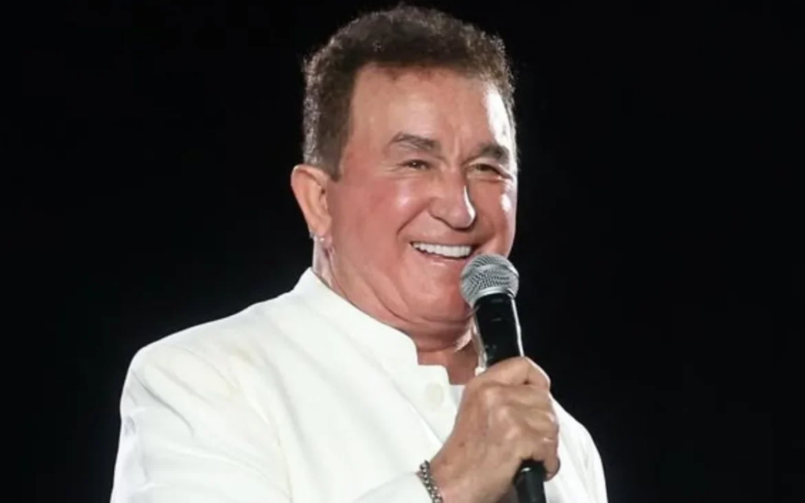 Cantor Amado Batista, de 75 anos, vestido de branco, sorrindo enquanto segura microfone em apresentação com fundo escuro