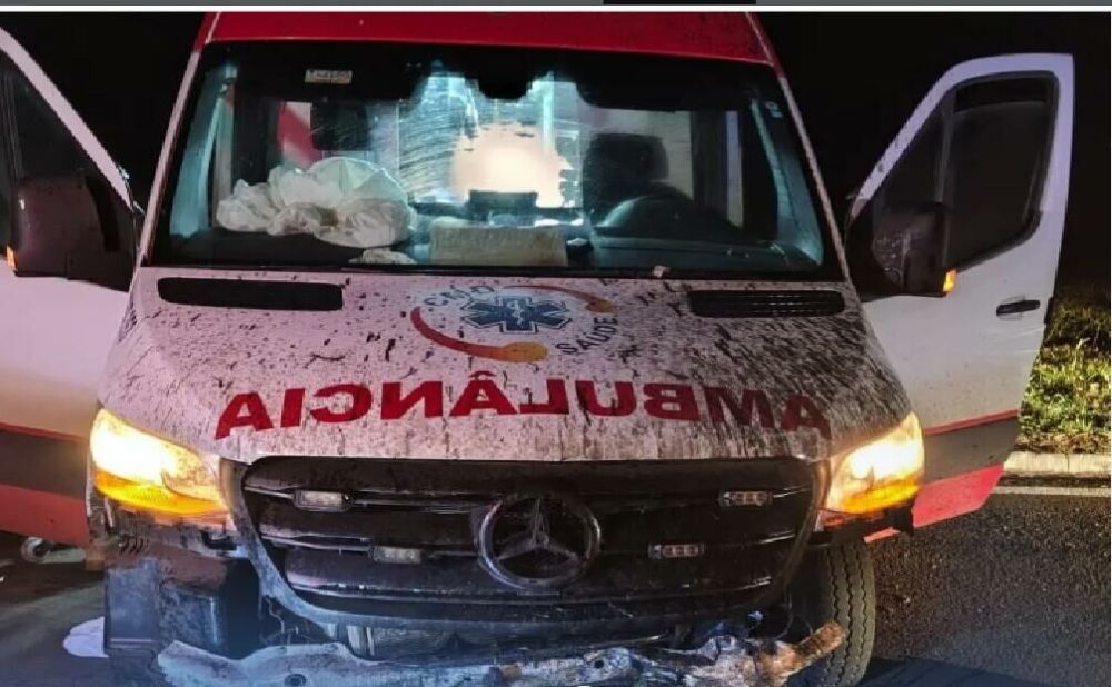 Motorista dorme ao volante e provoca acidente com ambulância de gestante intubada 6 Visão frontal de uma ambulância branca com a frente destruída e suja de terra após sair da pista na BR-262. O veículo está com as portas abertas e os faróis acesos durante a noite.