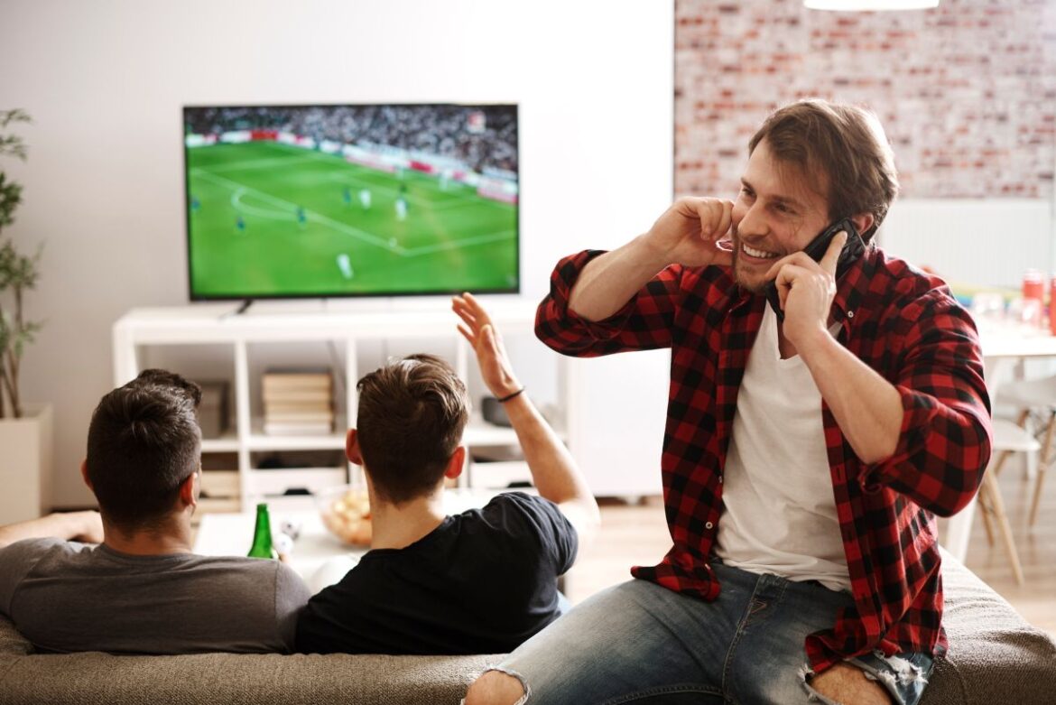 Três amigos em uma sala de estar assistindo a uma partida de futebol em uma televisão de tela plana; um deles sorri ao celular enquanto os outros gesticulam para a tela.