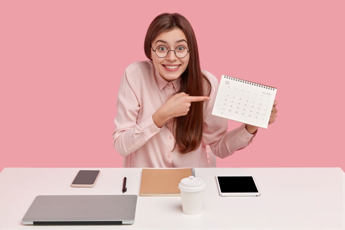 Uma mulher sorridente com óculos aponta para um calendário de mesa em um escritório, ilustrando o planejamento de feriados restantes do ano.