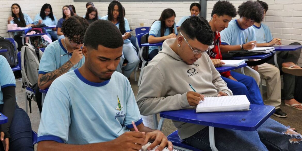 Grupo de jovens estudantes adultos assistindo aula e fazendo anotações em uma sala de aula de escola estadual.