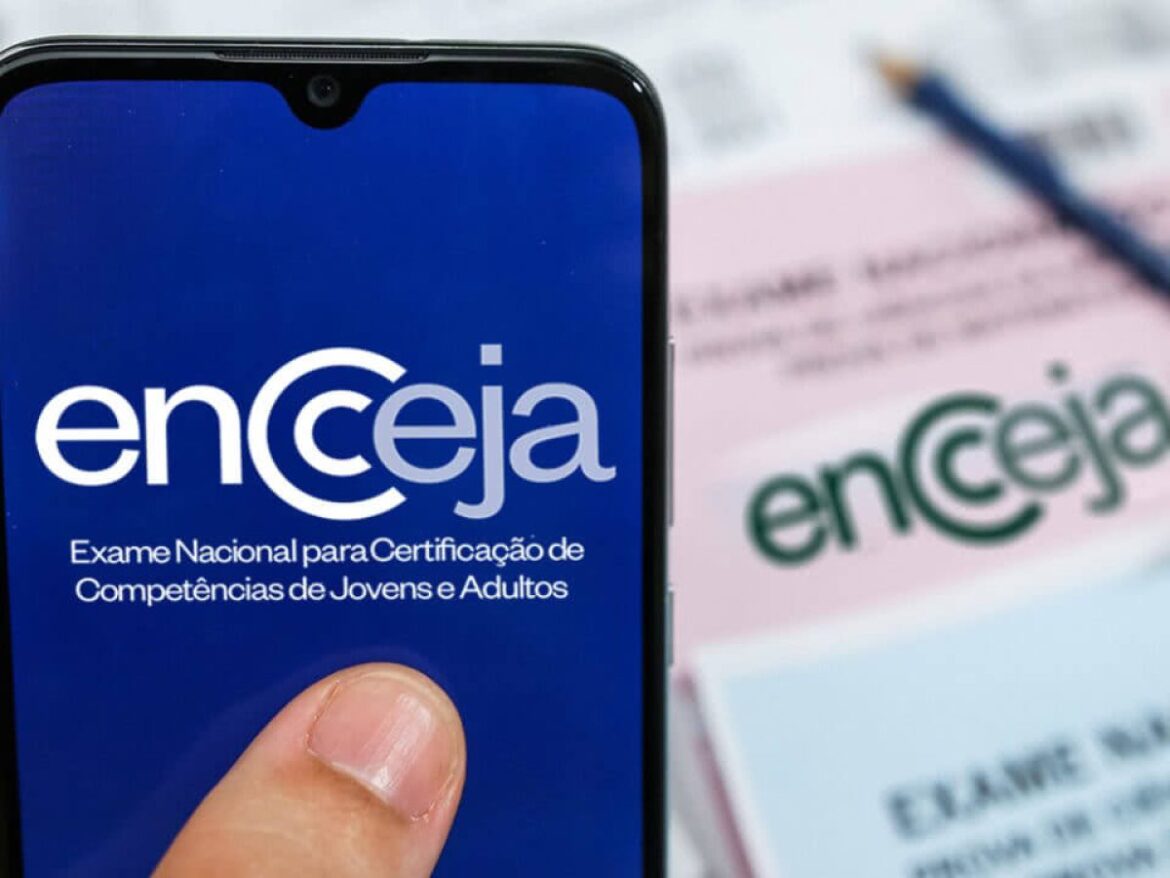 Close-up de um dedo indicador pressionando a tela de um celular que exibe o logo oficial do Encceja em branco sobre fundo azul, com material impresso ao fundo.