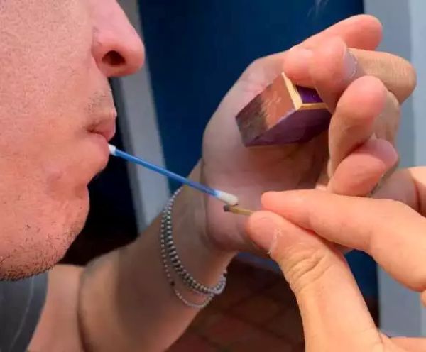 Fumar cotonete vira tendência entre jovens; saiba os perigos envolvidos 6 Close-up de um jovem segurando um cotonete de haste azul na boca enquanto outra pessoa aproxima um palito de fósforo aceso para queimar a ponta de algodão.