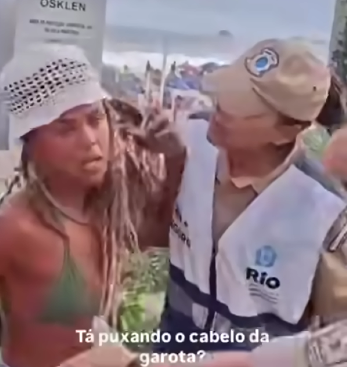 Vídeo de abordagem a artesã em Ipanema gera debate sobre fiscalização e direitos de trabalhadores informais 2 Frame de vídeo mostrando uma agente da Guarda Municipal do Rio de Janeiro segurando o cabelo de uma mulher loira de biquíni e chapéu branco durante uma abordagem em Ipanema. Abaixo, a legenda do vídeo diz: