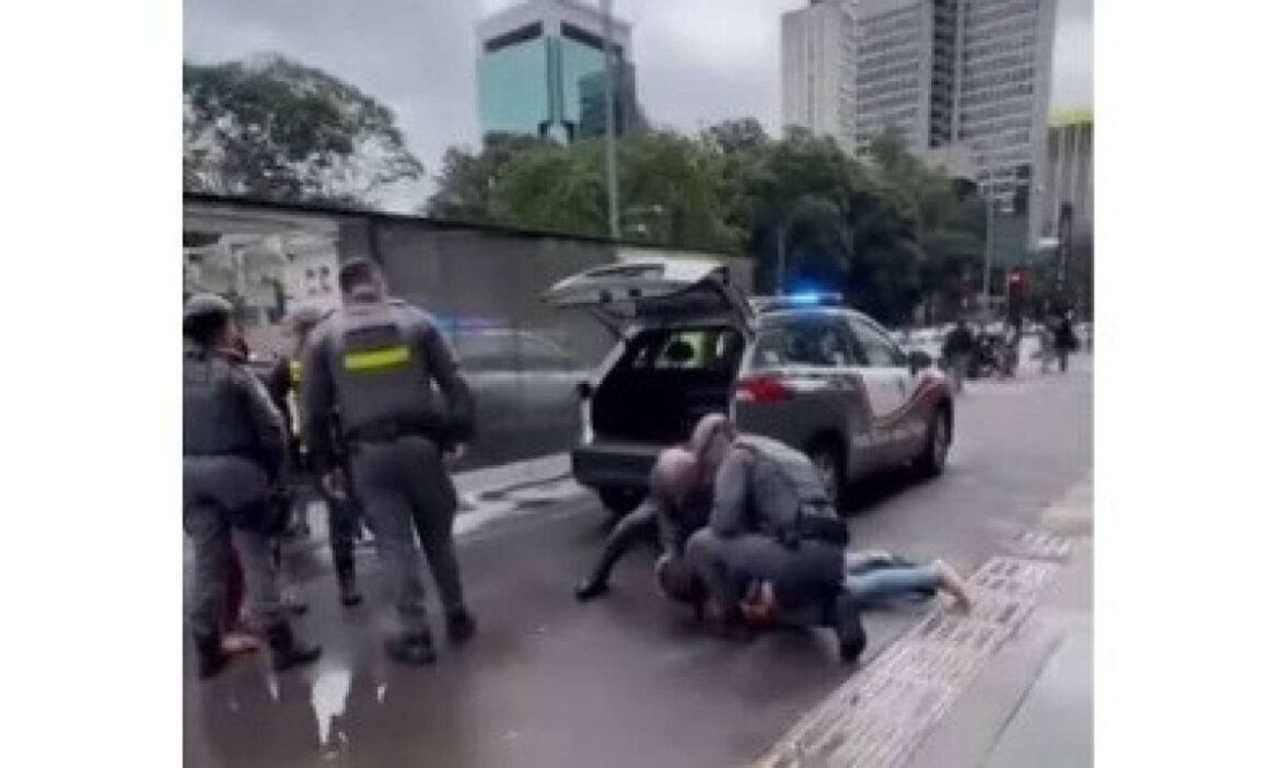 Frame de vídeo mostrando policiais militares na Avenida Paulista. Um policial imobiliza uma pessoa no chão, próximo à traseira de uma viatura com o porta-malas aberto e luzes de emergência acesas. Outros policiais e pedestres estão próximos. Prédios e árvores compõem o fundo sob um céu nublado.