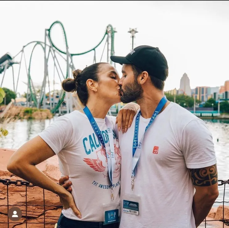 Daniel Cady, ex-marido de Ivete Sangalo, faz revelações surpreendentes sobre o término do casamento 6 Ivete Sangalo e Daniel Cady se beijando em um parque de diversões. Ivete está à esquerda, com o cabelo preso em um coque, vestindo uma camiseta branca com estampa e o braço esquerdo na cintura. Daniel está à direita, usando uma camiseta branca, boné preto e exibe uma tatuagem tribal no braço esquerdo. Ambos usam credenciais azuis penduradas no pescoço. Ao fundo, vê-se uma montanha-russa verde, um lago e prédios sob um céu claro.