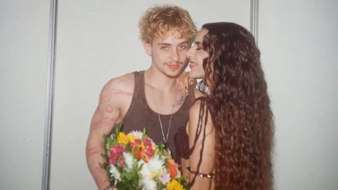 Marina Sena, com longos cabelos ondulados, abraçando Juliano Floss, que segura um buquê de flores, posando juntos em um ambiente de camarim.