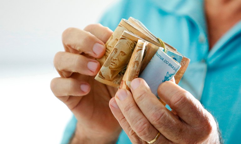 Governo do Brasil propõe novo salário mínimo para 2027; veja a novidade 6 Close-up das mãos de um homem contando várias notas de Real (50 e 100 reais) dobradas e presas por um elástico. O fundo mostra uma pessoa vestindo camisa azul clara.