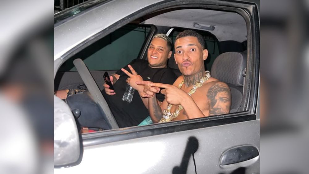 Justiça Federal decreta nova prisão de MC Ryan SP e MC Poze 6 MC Ryan SP e MC Poze do Rodo sorrindo dentro de um carro cinza. Ryan está no banco do motorista de camiseta preta e Poze está sem camisa no banco do passageiro, exibindo tatuagens e correntes de ouro.