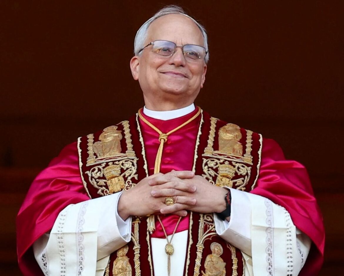 O Papa Leão XIV sorrindo ligeiramente e olhando para cima, com as mãos entrelaçadas, vestindo uma mozeta de veludo vermelho e dourado sobre um rócquete branco.