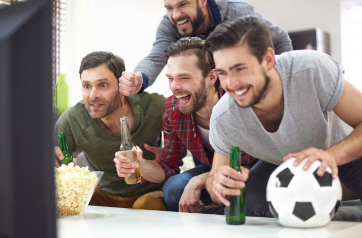 Copa do Mundo 2026 será exibida em TV 3.0 com qualidade 4K e som de cinema 6 Quatro homens jovens em uma sala de estar, vibrando e comemorando enquanto assistem a um jogo de futebol na televisão. Eles seguram garrafas de bebida, pipoca e uma bola de futebol, demonstrando entusiasmo.