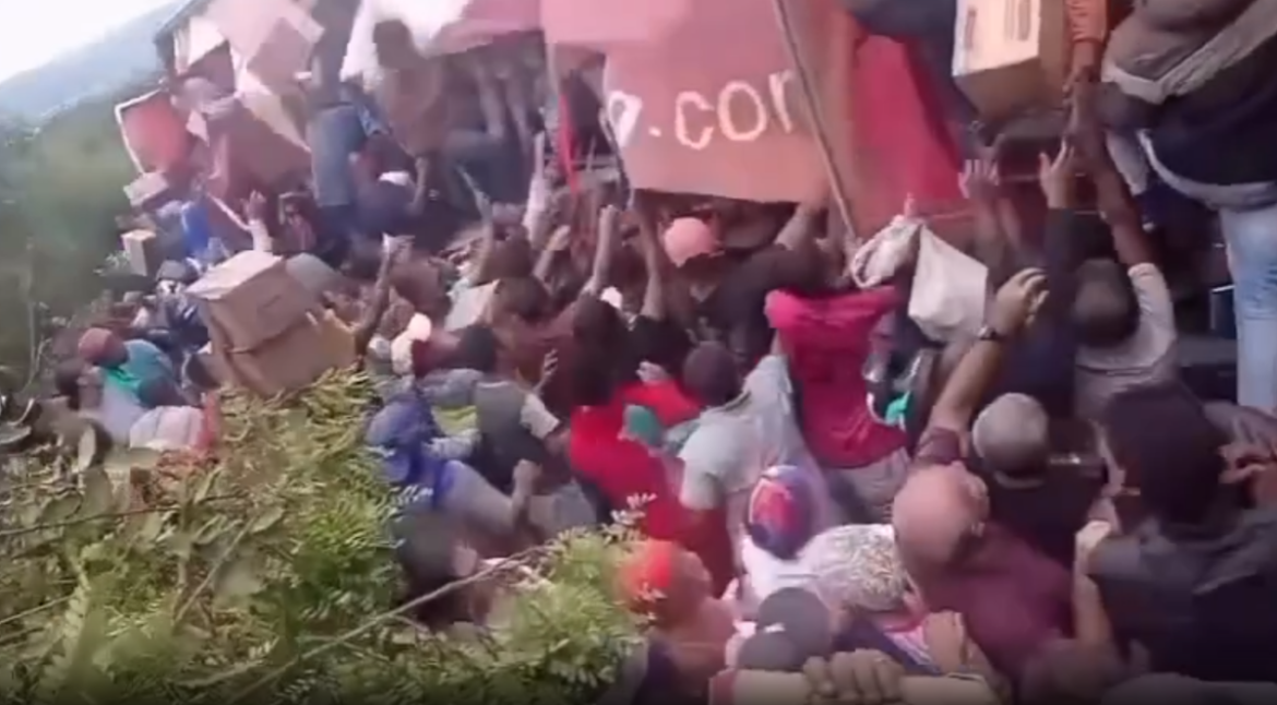 multidão aglomerada ao redor de uma carreta tombada na BR-116, na Bahia. Dezenas de pessoas esticam os braços para saquear caixas de leite em pó sobre a vegetação.
