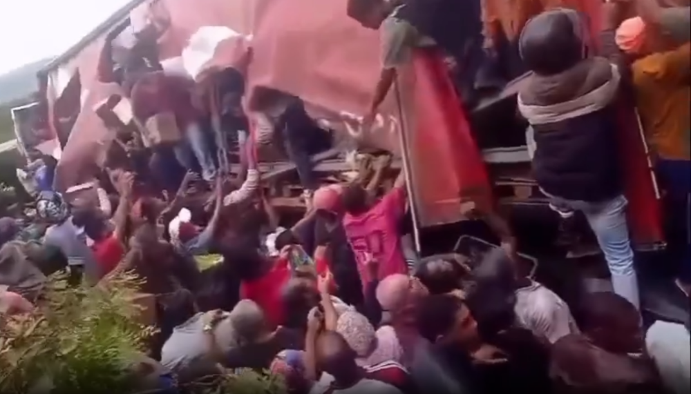 Imagens fortes: Carreta tomba e população saqueia toda a carga de leite em pó 6 Multidão de pessoas aglomerada ao redor de uma carreta de carga acidentada na vegetação, retirando caixas de papelão e sacos, ilustrando uma cena de saque após tombamento.