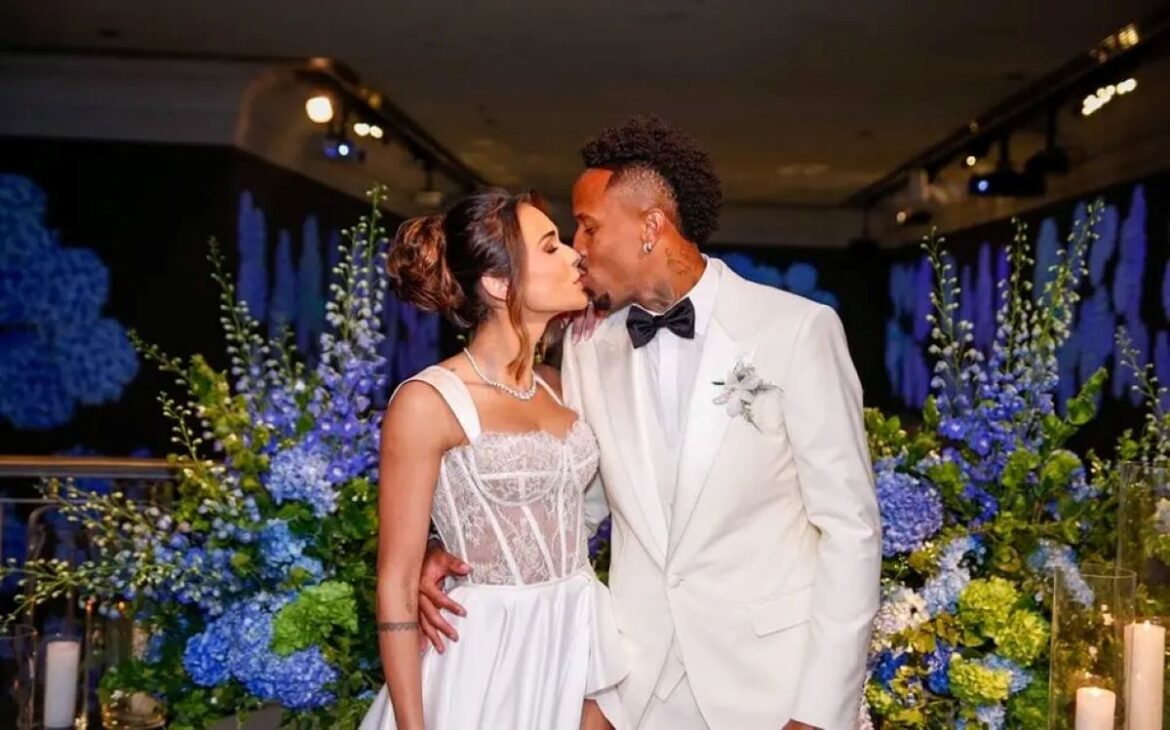 Tainá Castro e Éder Militão se beijam durante cerimônia de casamento. Ela usa vestido branco com corpete de renda e ele terno branco com gravata borboleta preta.