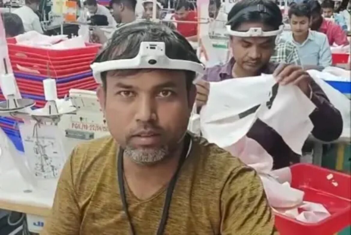 Rosto de um trabalhador têxtil indiano em primeiro plano, usando um dispositivo branco com câmera acoplada na testa. Ao fundo, outros trabalhadores aparecem com o mesmo equipamento, focados em tarefas manuais, como costura, em uma fábrica cheia.