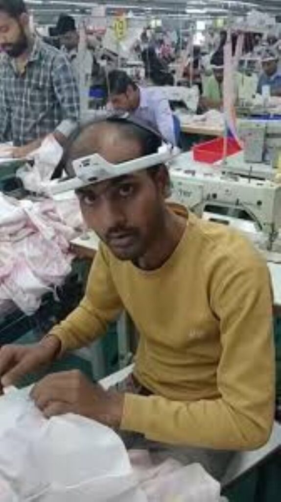 Vídeo: Trabalhadores usam câmeras na cabeça para treinar IA dos robôs que vão substituí-los 6 Foto de um trabalhador têxtil indiano em primeiro plano, usando um dispositivo branco com uma câmera acoplada à testa. Ele manipula tecidos claros em uma máquina de costura industrial, enquanto outros trabalhadores com o mesmo equipamento aparecem desfocados ao fundo em uma fábrica cheia.