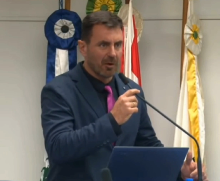 Vereador em vídeo polêmico defende leis contra mulheres trans em concursos 6 Um homem de meia-idade, com cabelos curtos e barba, usando um terno azul-marinho e uma gravata fúcsia, fala ao microfone em uma tribuna parlamentar, segurando a base do microfone com a mão direita. Ele está olhando para a direita. Ao fundo, há bandeiras desfocadas e fitas azuis e brancas com flores amarelas, sugerindo um ambiente da Câmara Municipal. Um notebook azul está sobre a tribuna à direita.