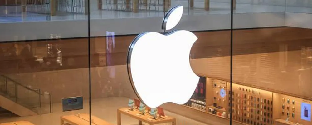 Apple pode pagar até R$ 467 a donos de iPhone; veja se você tem direito 6 Fachada de vidro da Apple Store com logo branco iluminado e produtos expostos em mesas de madeira no interior