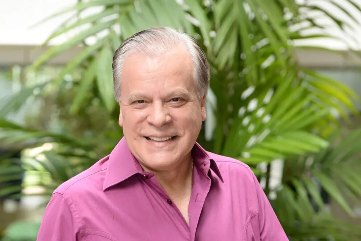 Chico Pinheiro sorrindo, cabelos grisalhos, camisa rosa magenta, fundo com vegetação tropical verde