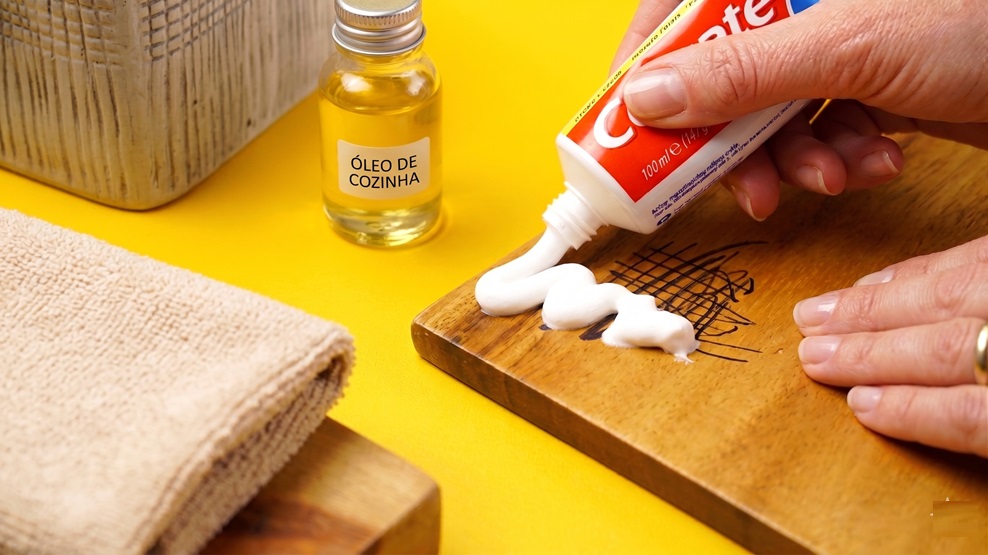 Como tirar risco de caneta do móvel de madeira? O produto do banheiro que salva a pintura 6 Mãos aplicando pasta de dente branca sobre rabiscos de caneta em tábua de madeira, com óleo de cozinha e pano bege ao lado
