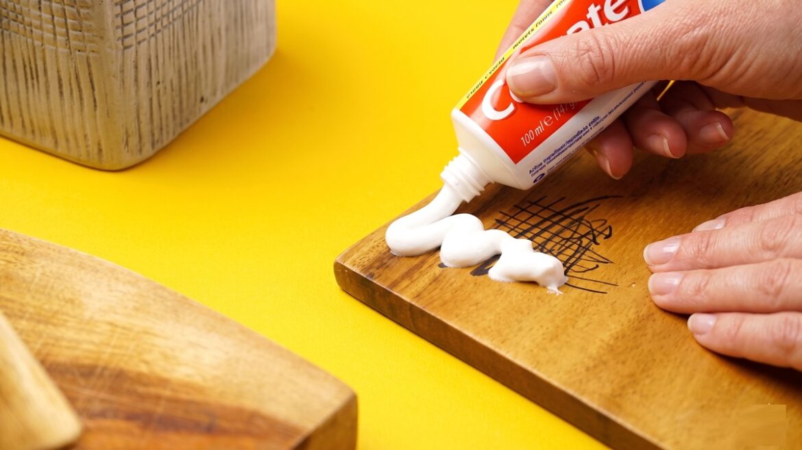 Mão aplica pasta de dente branca sobre riscos de caneta em tábua de madeira com fundo amarelo