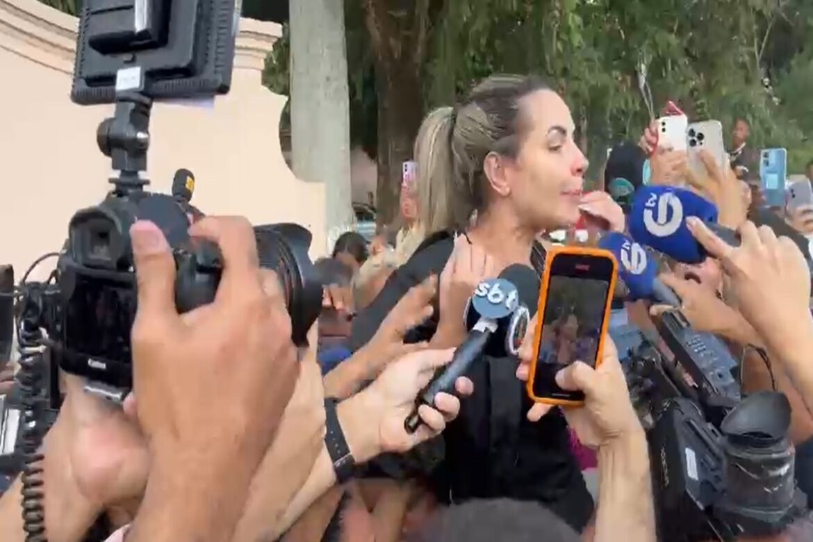 Deolane Bezerra de cabelo preso e blusa preta, rodeada por microfones do SBT e celulares de jornalistas em área externa