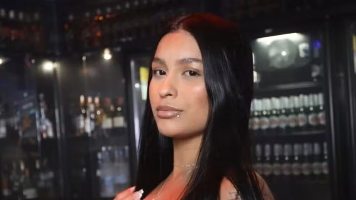 Mulher jovem de cabelos longos, pretos e lisos em ambiente de bar, com piercing no lábio inferior e expressão tranquila
