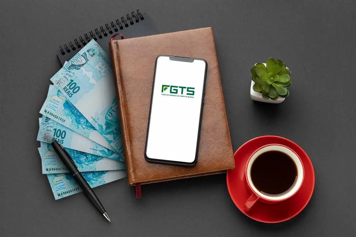 Governo libera FGTS para quem aderiu ao saque-aniversário e perdeu o emprego 6 Smartphone exibindo logo FGTS sobre agenda marrom, notas de R$ 100, caneta preta, xícara vermelha de café e suculenta