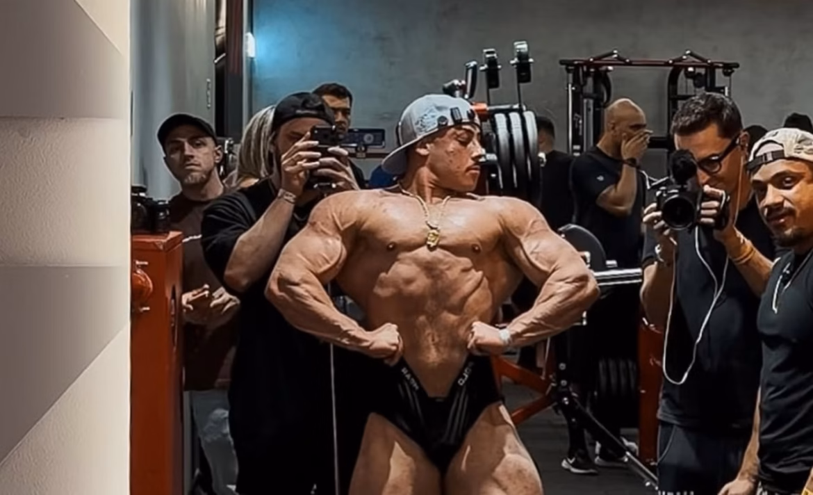 Fisiculturista Gabriel Ganley, de boné cinza e corrente dourada, posa em academia enquanto pessoas ao redor filmam