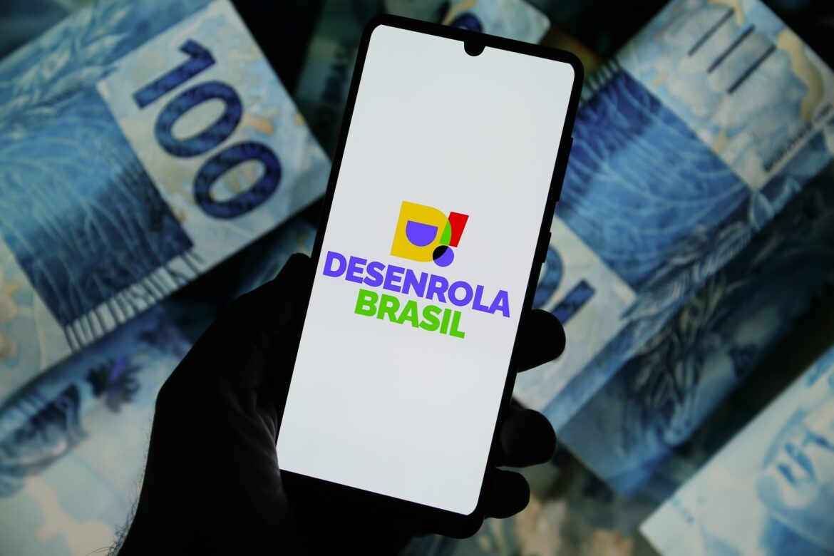 Mão segurando celular com logo do Desenrola Brasil sobre notas de R$ 100 espalhadas ao fundo