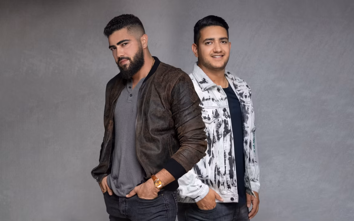 Henrique e Juliano posam lado a lado com fundo cinza; Henrique usa jaqueta marrom e Juliano veste jaqueta branca tie-dye