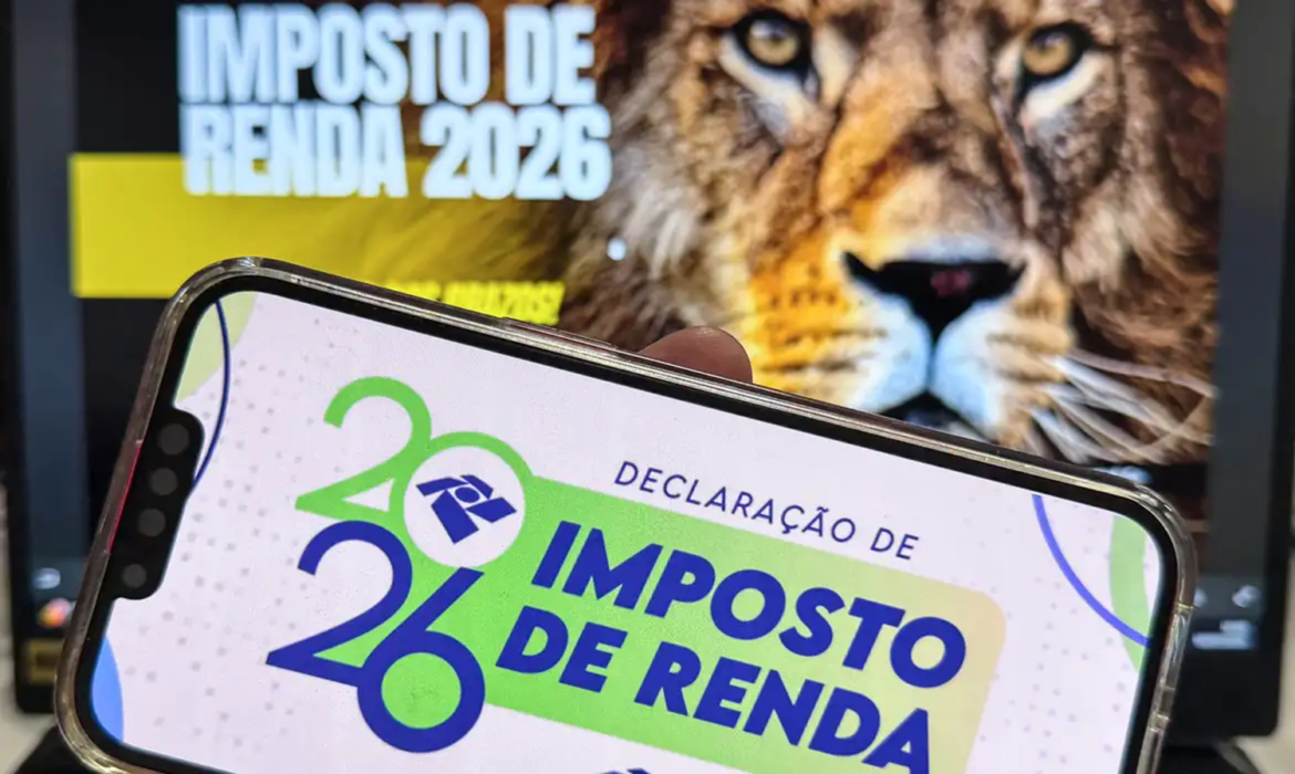 Celular exibindo tela do programa Imposto de Renda 2026 da Receita Federal, com leão em monitor ao fundo