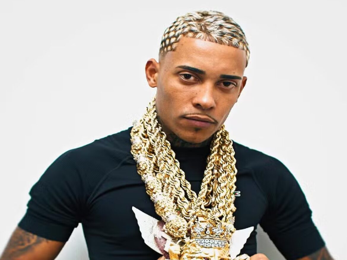 MC Poze do Rodo de camisa preta com tranças loiras e grossas correntes de ouro no pescoço, pingente de coroa cravejado