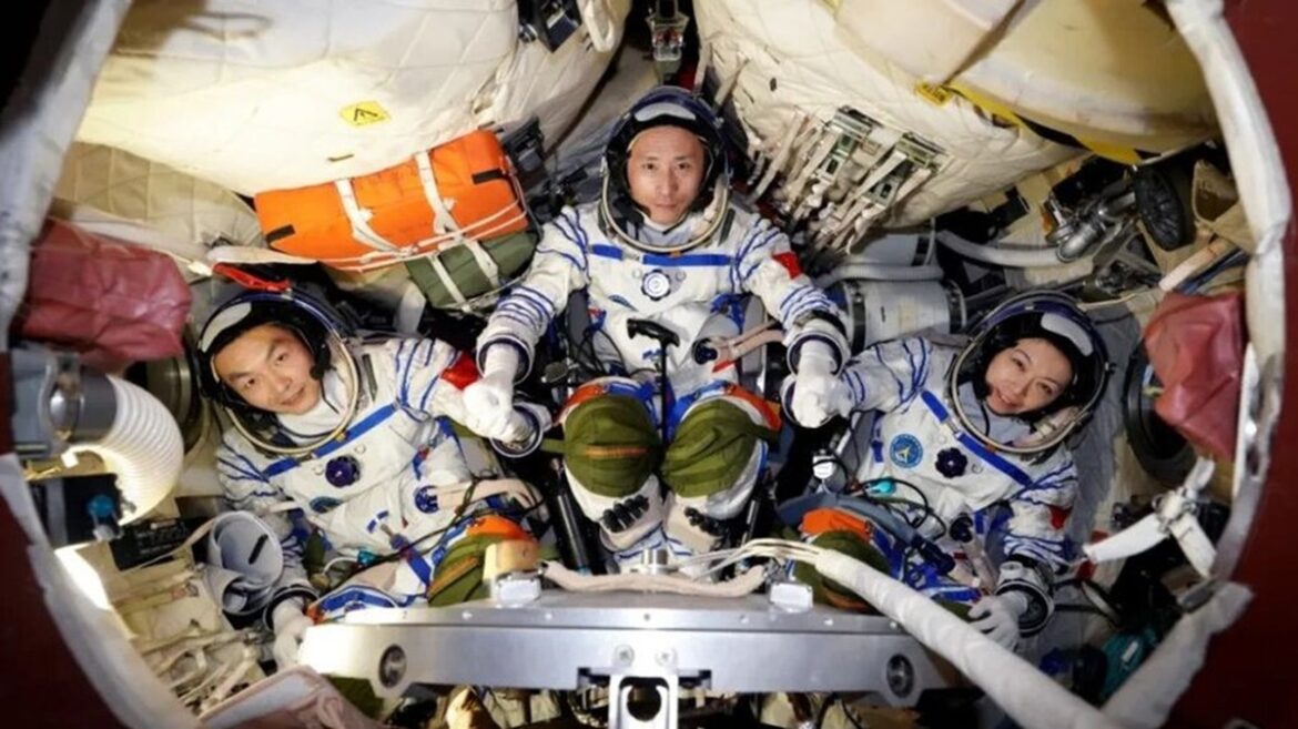 Astronautas Zhu Yangzhu, Zhang Zhiyuan e Lai Ka-ying em trajes espaciais brancos com detalhes azuis e laranja