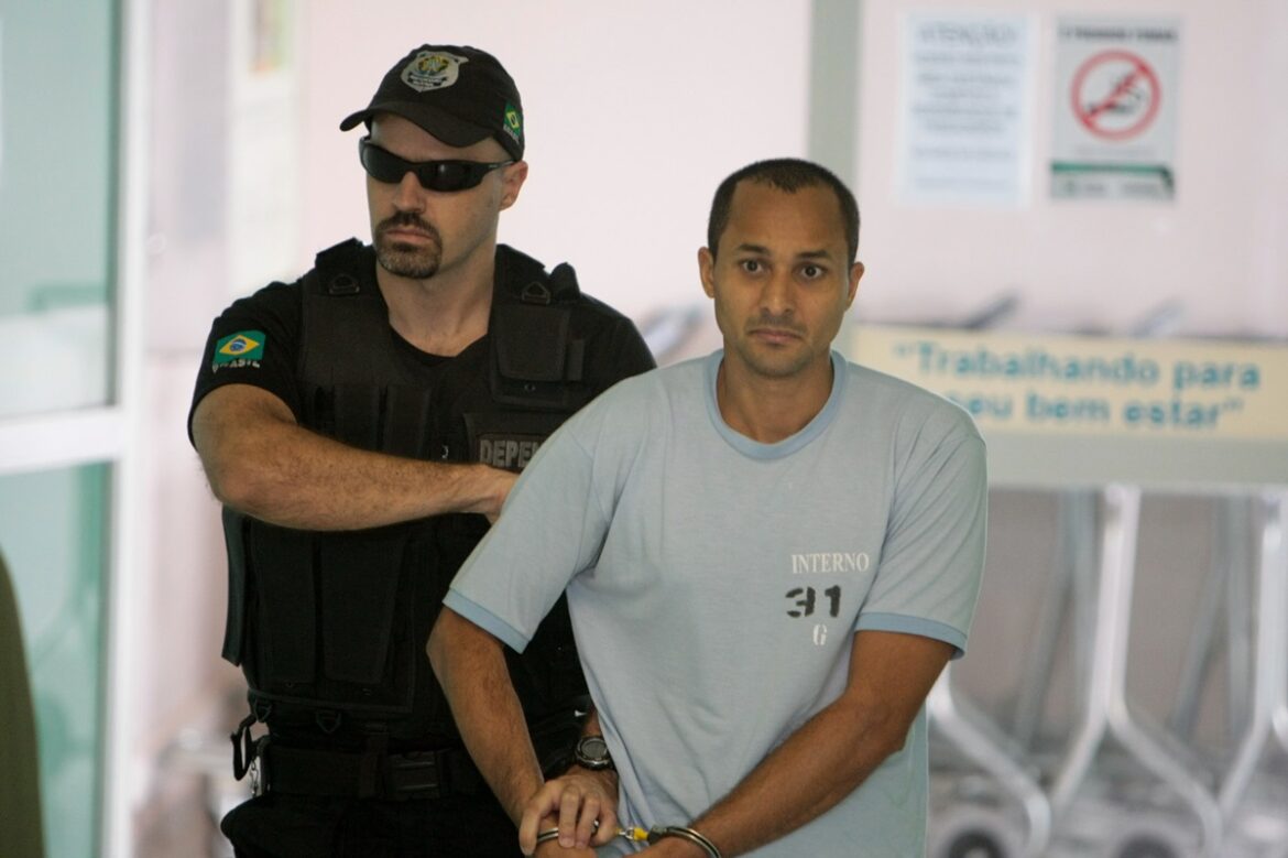 Preso algemado com camiseta azul "Interno 31" conduzido por agente do DEPEN fardado de preto em ambiente prisional