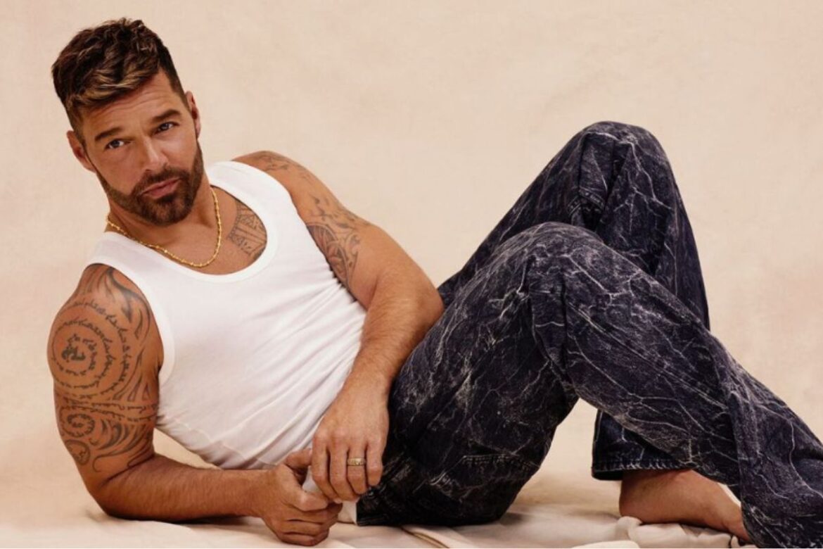Ricky Martin tatuado usando regata branca e calça jeans marmorizada, deitado em pose casual com corrente dourada