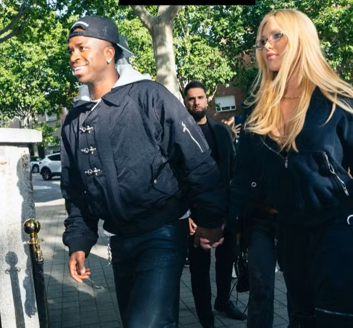 Casal caminha de mãos dadas em rua arborizada, ele com jaqueta preta e boné, ela loira com óculos e jaqueta escura