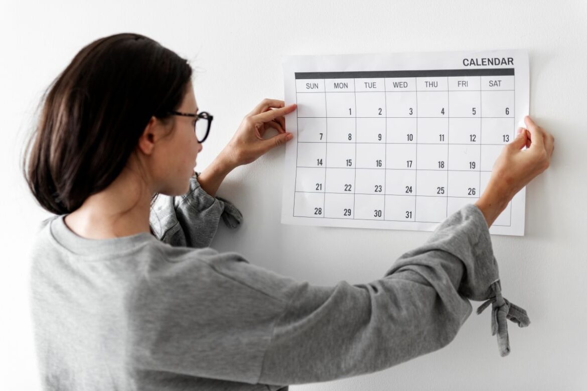 Uma mulher de óculos e blusa cinza segura um calendário de papel branco contra uma parede clara, focando no planejamento de datas.