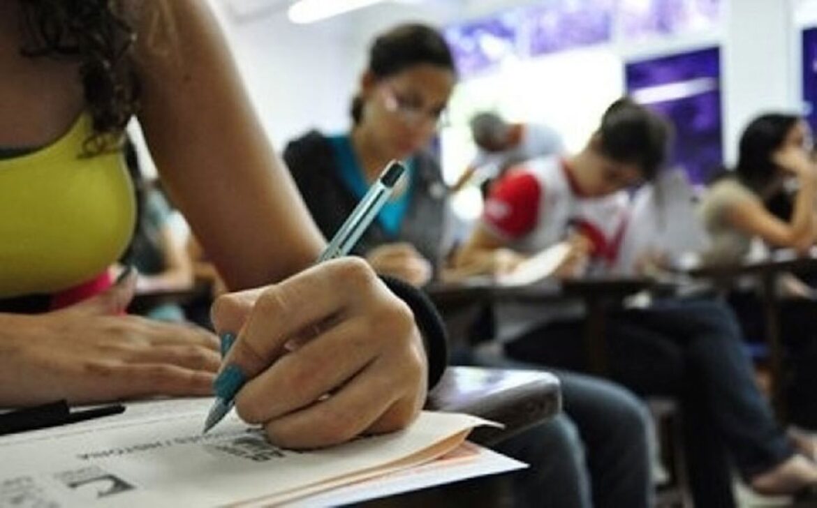 Mão de uma estudante escreve durante uma prova e, ao redor, outros candidatos sentados nas carteiras