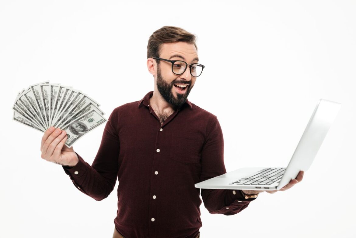 Um homem sorridente, usando óculos e barba, segura um notebook aberto em uma das mãos e um leque de notas de dólar na outra.