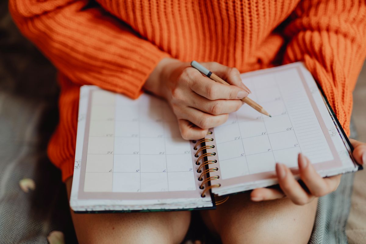 Faltam só seis feriados em 2026; veja quando vão acontecer 6 Close das mãos de uma pessoa usando uma blusa de tricô laranja, segurando um lápis e escrevendo em uma agenda aberta com layout de calendário mensal.