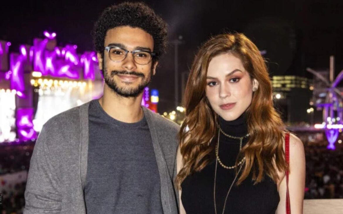 Sérgio Malheiros de óculos e cardigan cinza ao lado de Sophia Abrahão de blusa preta em evento noturno com luzes roxas