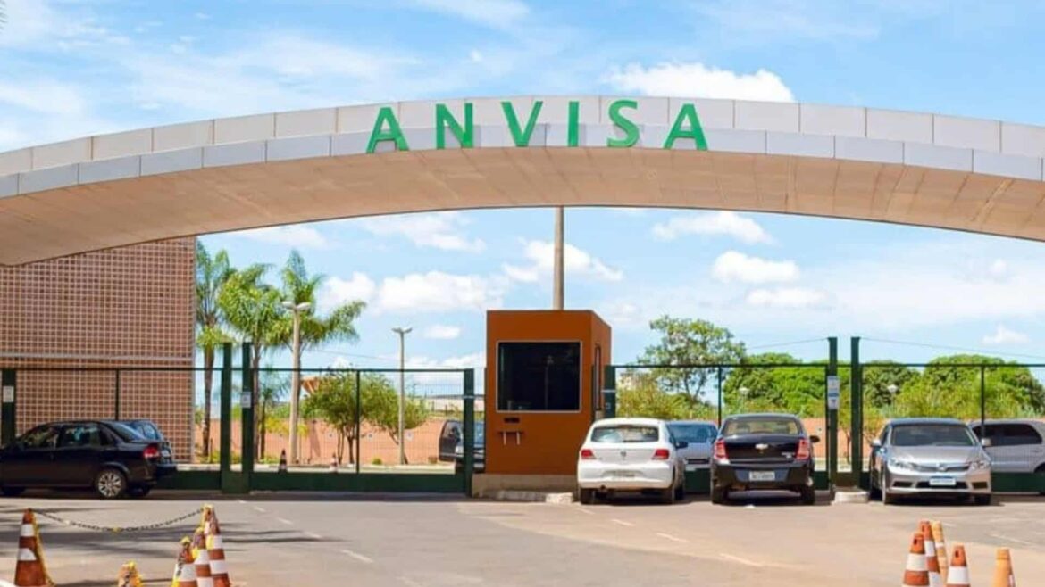Fachada da sede da Anvisa com arco cinza e letreiro verde, portaria com guarita laranja, carros estacionados e cones de sinalização